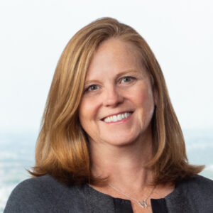 Close cropped professional photo of a red-haired woman, Kathleen Garrity, smiling and wearing a charcoal and black blazer cardigan over a black blouse with an out of focus light blue background.