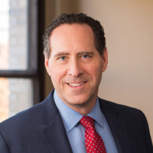 Professional up close photo of a man, Steven M. Williams, with short, curly, brown hair, smiling into the camera, standing in a tan office wearing a navy suit with a light blue button down shirt and a red tie.