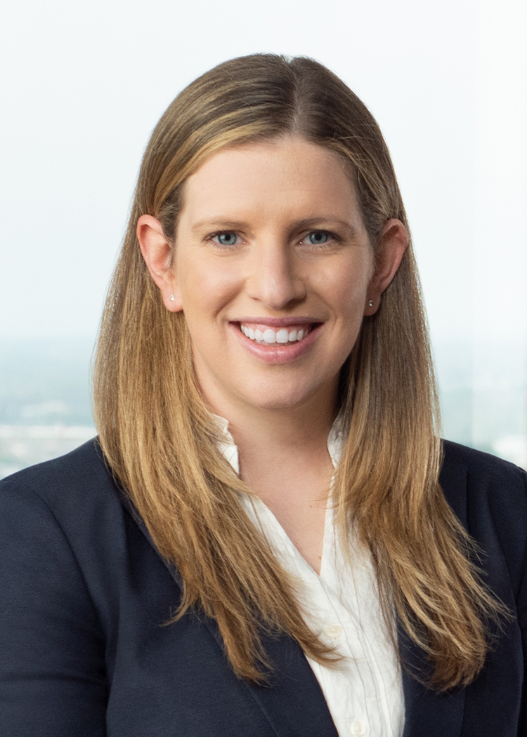 close crop professional headshot of Dana Hasness in a blue suit and white blouse standing in front of a wall of windows and an orange couch