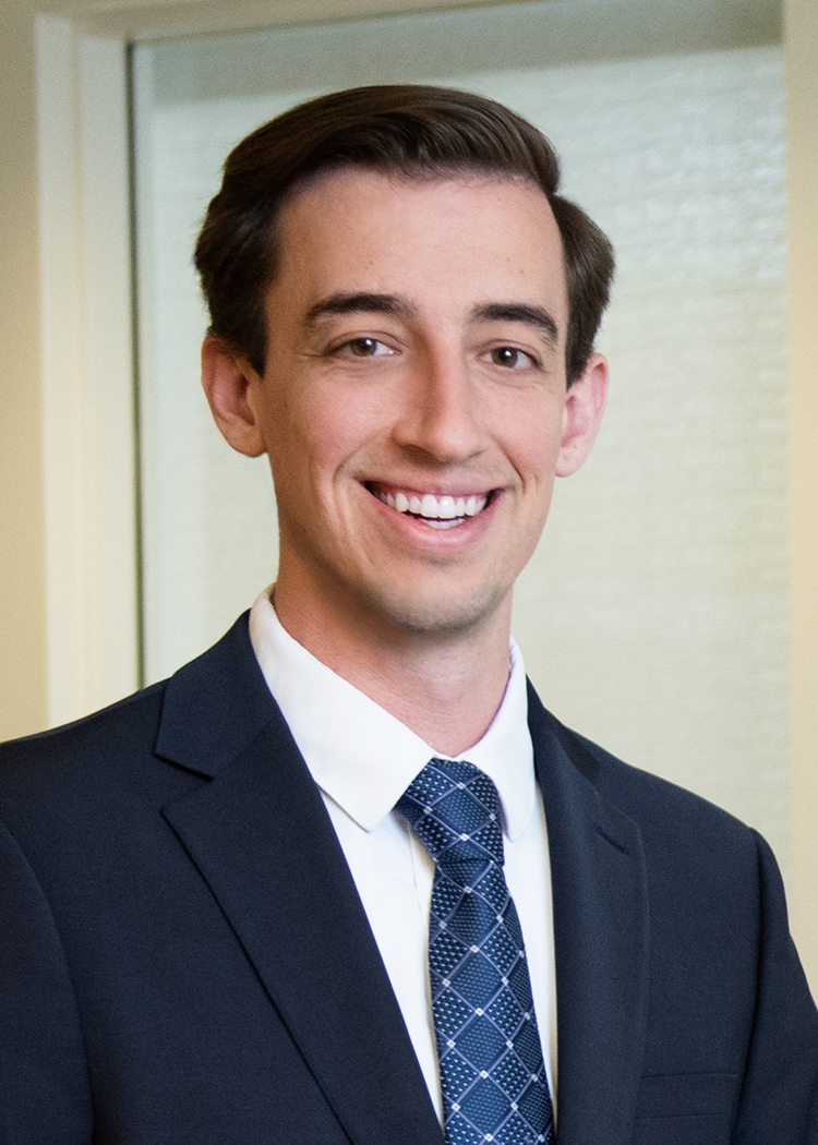 Headshot of Nicholas Bencsics, standing in an office, wearing a dark suit and a blue suit