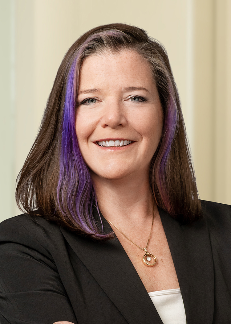 Close-cropped professional photo of Carolyn Daly smiling with shoulder length, straight brown hair with a purple streak wearing a black blazer with a white shirt with an off-white background.