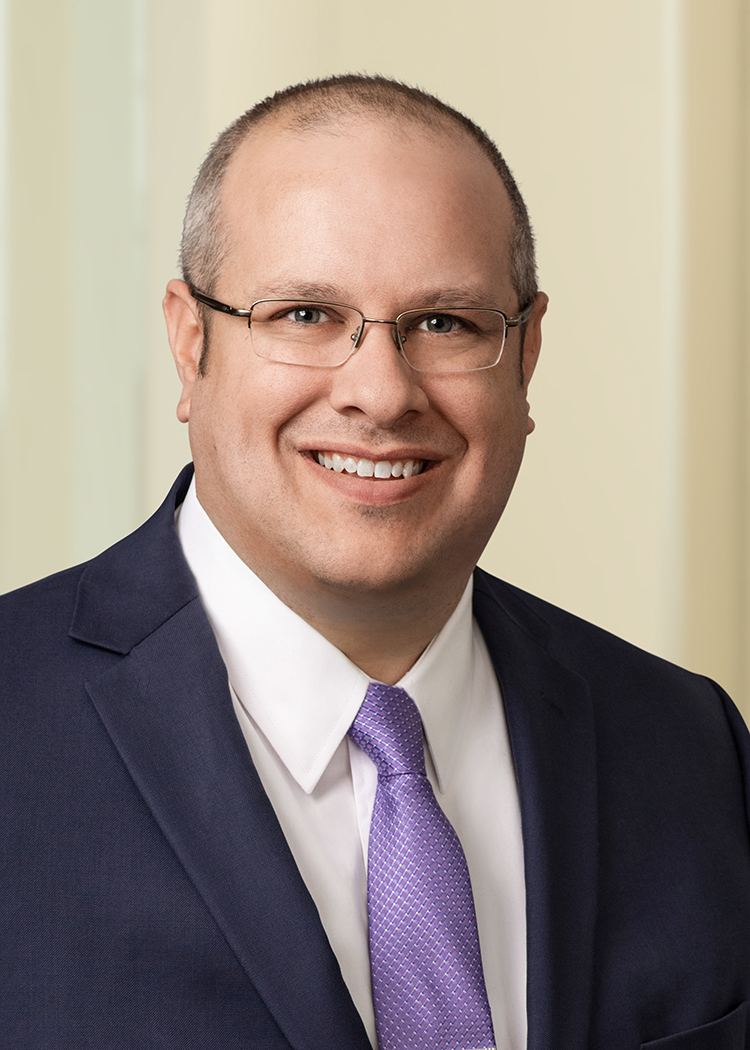 Close-cropped professional photo of Sean Gaynor smiling with frameless glasses wearing a navy suit with a white shirt and light purple tie with an off-white background.