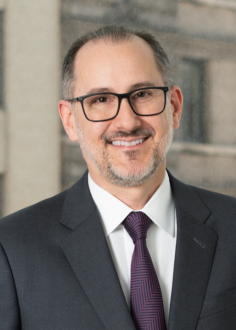 Close up professional photo of Paul Lucas smiling and wearing black rectangular glasses, a charcoal suit white button up, and a maroon striped tie with a window overlooking Wall Street as the background.