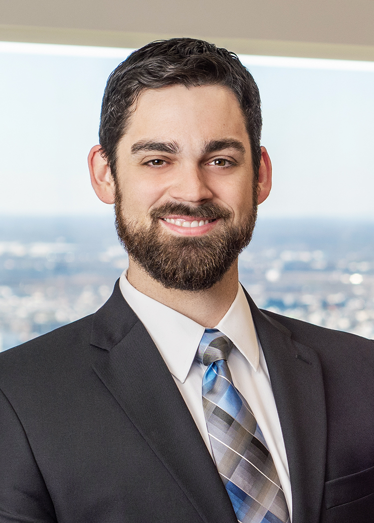 Close-cropped professional photo of a young man, Christopher Ouellette, smiling with a brown beard and wearing a charcoal suit, white collared shirt, and a blue and silver plaid tie in front a window overlooking Philadelphia.