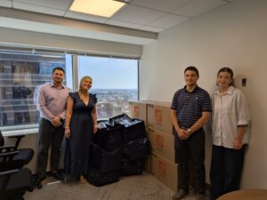 Four Cohen Seglias team members stand beside stacks of large boxes and blue donation bags filled with clothing collected during the firm's Spring Clothing Drive. The group smiles in a conference room overlooking the city skyline through large windows, celebrating the successful collection before the donations were delivered.